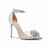 A single metallic silver high-heeled sandal with an ankle strap, clear side panel, and a rhinestone-embellished flower detail on the toe, shown against a plain white background.