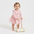 A smiling toddler girl in a pink long-sleeve dress with a tulle skirt pushes a small wooden toy walker on a clean white studio background.