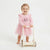 A smiling toddler stands barefoot on a white studio background, holding a small wooden push toy. The child wears a light pink long-sleeve dress with a tulle skirt and a sequined cat motif on the chest.