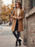 A woman stands on a city sidewalk beside a brick wall, wearing a camel-colored mid-length coat over a cream turtleneck, dark skinny jeans, and black ankle boots.