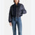 A model wearing a cropped navy blue bomber jacket with mixed textures (satin, corduroy, and ribbed knit) paired with blue jeans, shot against a light gray background.