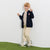 A young boy stands against a white wall wearing a navy blue zip-up cardigan over a beige and white striped shirt, paired with beige pants and green-and-white sneakers.