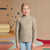 A young girl with blonde hair stands in a child's bedroom wearing a beige ribbed mock neck sweater and light blue jeans.