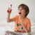 A young child sitting on a white bed wearing an orange t-shirt and patterned pajama pants, holding a red and white toy rocket up in the air with a surprised expression.