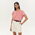 A woman modeling a dusty pink short-sleeve t-shirt tucked into cream-colored pleated shorts with a brown belt, against a plain white studio background.