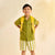 A young boy stands with hands on hips wearing a matching olive green short-sleeve button-up shirt and striped shorts, styled with a bright yellow backpack and green sandals against a warm cream backdrop.