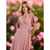 A woman wearing a dusty pink wrap-style midi dress with flutter sleeves and a tie waist, standing outdoors against a blurred background of pink flowers and green foliage.