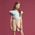 A young girl with long light brown hair stands against a solid dusty rose background, wearing a light blue ruffled-sleeve top and beige linen shorts.