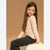 A young girl with long brown hair sits on a white textured cushion against a solid tan studio background. She is wearing a beige long-sleeve knit top with small black heart patterns and dark gray pants.