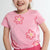 A young girl smiling and looking to the side, wearing a pink short-sleeve t-shirt with a pink and yellow flower pattern, paired with pink drawstring shorts against a light gray background.