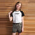 A young girl with long light brown hair stands in front of a wooden slat wall, wearing a raglan-style short-sleeve t-shirt with a white body and black sleeves, featuring a black graphic that reads 'HAWAIIAN BEACH'. She is also wearing gray denim shorts.