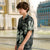 A young boy wearing a matching short-sleeve button-up shirt and shorts set with a black tropical leaf pattern, standing outdoors in front of a fountain and classical buildings.
