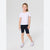 A young girl modeling a white short-sleeve crewneck t-shirt, dark navy bike shorts, and white low-top sneakers with red stripes, standing against a light gray studio background.