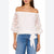 An off-the-shoulder white blouse with sheer lace sleeves and a white bow detail at the waist, worn by a model with blue jeans against a white background.