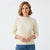 A woman wearing a cream-colored ribbed knit mock neck sweater and blue jeans, standing against a plain white studio background.