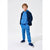 A young boy modeling a blue children's tracksuit consisting of a heather blue hoodie, matching blue sweatpants, and navy blue slip-on shoes, styled with a navy blue puffer jacket over his shoulders, against a plain light gray studio background.