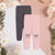 Two pairs of children's jogger sweatpants laid flat: one dark gray and one light pink, both with side pockets and bow graphics near the lower leg. The pink pair is in front, and the scene is styled with white flowers and greenery on a split white and blush background.