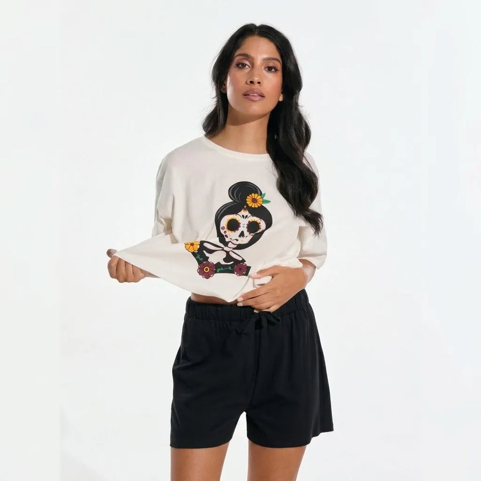 A woman modeling a cream-colored long-sleeve t-shirt with a colorful Day of the Dead sugar skull graphic, paired with black shorts, against a plain white studio background.