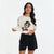A woman modeling a cream-colored long-sleeve t-shirt with a colorful Day of the Dead sugar skull graphic, paired with black shorts, against a plain white studio background.