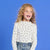 A young girl with curly blonde hair smiling and looking to the side, wearing a white long-sleeve top with black polka dots and blue jeans, set against a solid blue studio background.