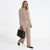 A woman models a beige tailored two-piece suit consisting of a cropped blazer and matching wide-leg trousers, paired with black flats and holding a black structured handbag against a plain white studio background.