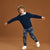 A young boy wearing a navy blue sweater and matching navy patterned pants with white sneakers, posing with outstretched arms against a solid brown studio background.