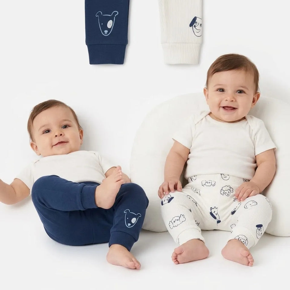 Two babies sitting on a white floor wearing matching white and navy blue baby clothing with cartoon bear prints, with two more matching garments hanging above them against a white background.