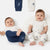 Two babies sitting on a white floor wearing matching white and navy blue baby clothing with cartoon bear prints, with two more matching garments hanging above them against a white background.