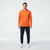 A man modeling a bright orange long-sleeve athletic hoodie with black pants and white sneakers, standing against a light gray studio background.