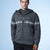 A man wearing a dark gray zip-up knit hooded sweater with a white Nordic snowflake pattern across the chest and sleeves, paired with a dark gray t-shirt and black pants, set against a light blue studio background.