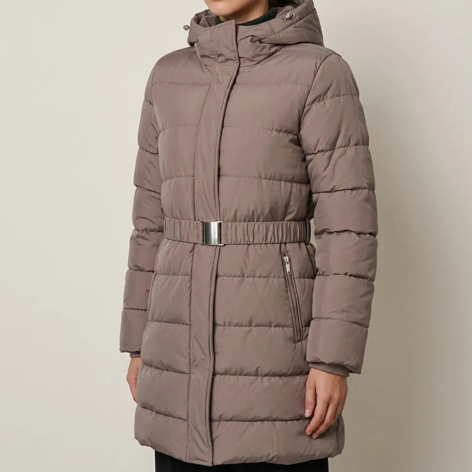 A woman wearing a mid-length taupe puffer coat with a hood, zippered front, and a matching elasticated belt with a silver buckle, shown against a plain light gray background.