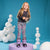 A young girl with long blonde hair stands on a lavender platform wearing patterned leggings with a pink, black, and white leopard print, a dark gray long-sleeve shirt, and purple ski boots. The background is a bright blue studio backdrop decorated with white stacked foam spheres.