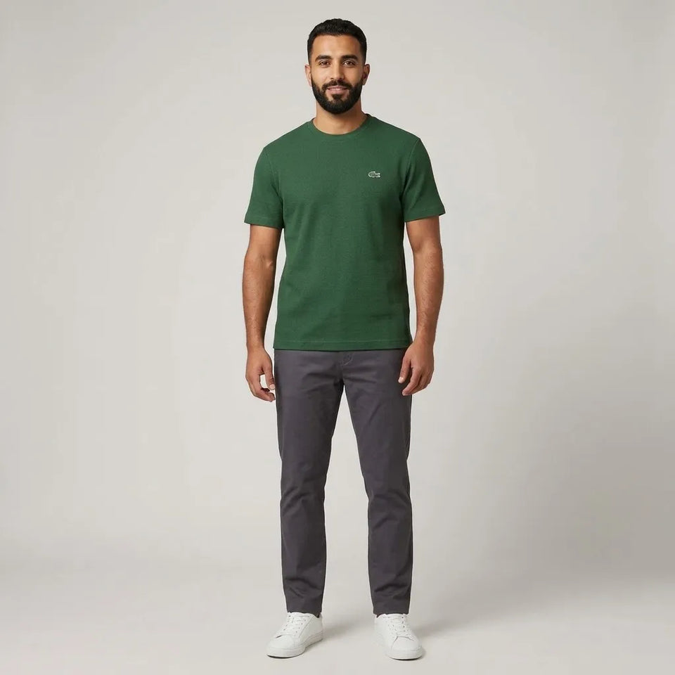 A man modeling a green short-sleeve crewneck t-shirt with a small white Lacoste crocodile logo on the left chest, paired with dark gray trousers and white sneakers, against a light gray studio background.