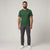 A man modeling a green short-sleeve crewneck t-shirt with a small white Lacoste crocodile logo on the left chest, paired with dark gray trousers and white sneakers, against a light gray studio background.