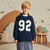 A young boy with blonde hair stands in a child's bedroom wearing a navy blue long-sleeve sweatshirt with the number '92' printed in white on the front, paired with blue jeans.