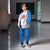A child model wearing a two-piece tracksuit consisting of a light gray and blue zip-up hoodie with a basketball graphic, and matching blue jogger pants, standing in a school hallway.
