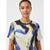 A woman wearing a short-sleeve crewneck t-shirt with an abstract watercolor print in blue, purple, yellow, and cream, set against a plain light gray background.