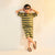 A child wearing a matching two-piece striped lounge set with a short-sleeve top and shorts in olive green and yellow, styled with dark sunglasses and brown sandals against a plain light background.