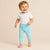 A smiling toddler standing barefoot against a warm beige studio background, wearing a plain white short-sleeve top and light aqua pants.