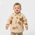 A smiling toddler wearing a cream-colored fleece button-up cardigan with brown teddy bear appliques and a matching beige pair of pants, photographed against a solid light gray background.
