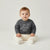 A baby sitting cross-legged on a light gray studio background, wearing a dark gray long-sleeve sweatshirt with a white dinosaur graphic and cream pants.