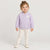 A young child stands against a light gray studio background wearing a lavender zip-front jacket with a small white flower applique on the chest, cream pants, and white shoes.