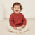 A smiling toddler sitting cross-legged on a cream carpet, wearing a textured rust-red knit sweater and cream ribbed leggings.