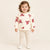 A toddler girl stands against a warm off-white studio background wearing a cream sweatshirt with red ribbon patterns, matching cream leggings, and white shoes.