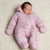 A sleeping newborn baby wearing a pink floral print hooded footed onesie, lying on a soft cream-colored textured blanket.