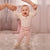 A baby wearing a cream long-sleeve top and light pink pants with a side bow, smiling while holding an adult's hands to learn to walk on a fluffy white rug.