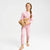 A young girl with a braided ponytail stands barefoot against a light gray studio background, wearing a pink long-sleeve pajama set with small darker pink polka dots and holding a brown teddy bear.