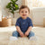 A smiling baby sitting on a fluffy cream rug wearing a navy blue ribbed short-sleeve t-shirt with a white Nike swoosh, blue jeans, and white socks in a bright, warm room.