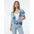 A woman modeling a short-sleeve, open-front tie-front cardigan with a blue watercolor floral print over a white tank top and light-wash jeans, set against a plain white studio background.
