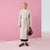 A woman models a long, light beige wrap coat with a tie waist, paired with dark blue cropped pants, brown sneakers, and a brown handbag, set against a pink and white studio backdrop.